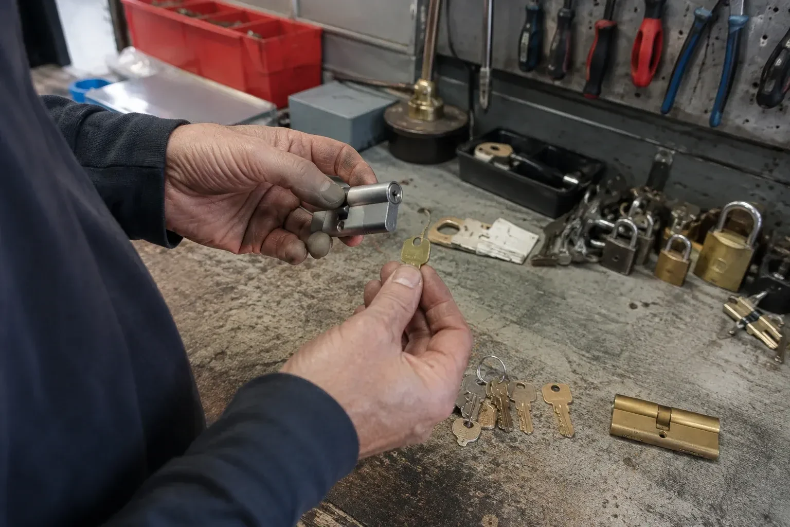 Hands holding a silver-colored lock cylinder and a brass-colored key above a workbench with padlocks, keys and tools. — Master Key System Design, Whitby